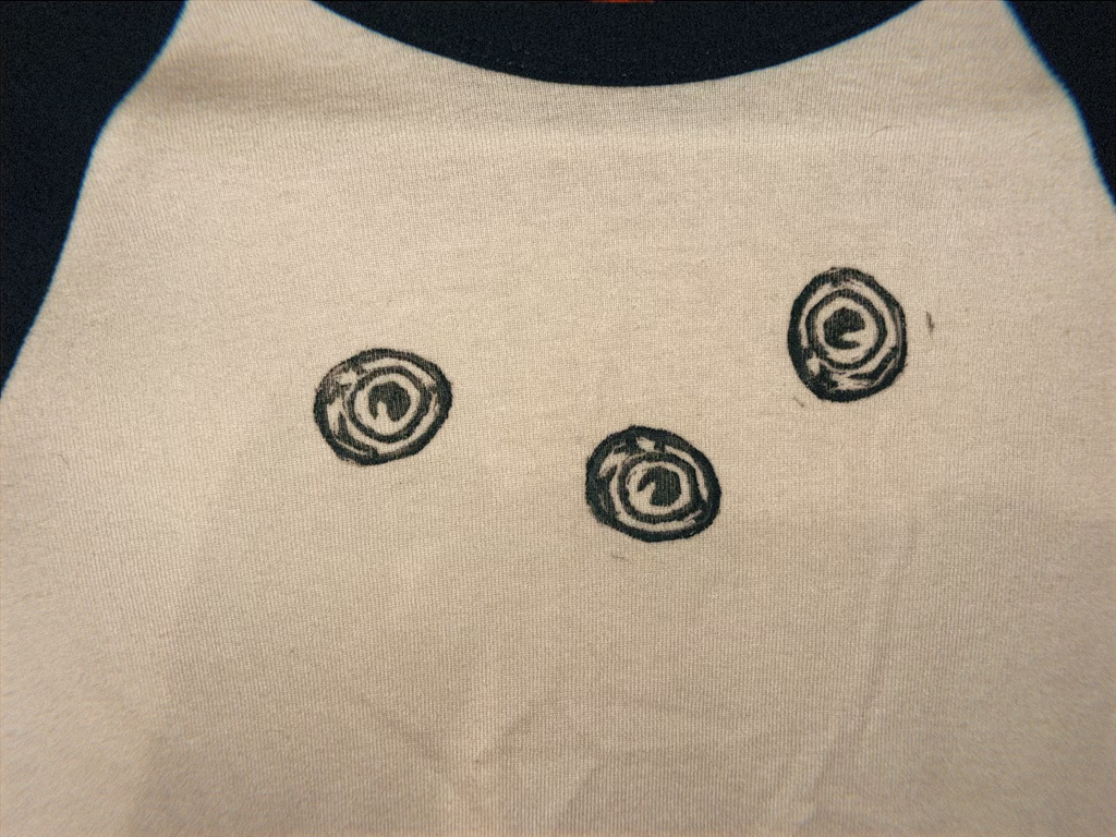 A close-up of a light beige fabric surface with three small black spiral or eye-like printed motifs arranged vertically. A dark curved shape appears along the right edge.