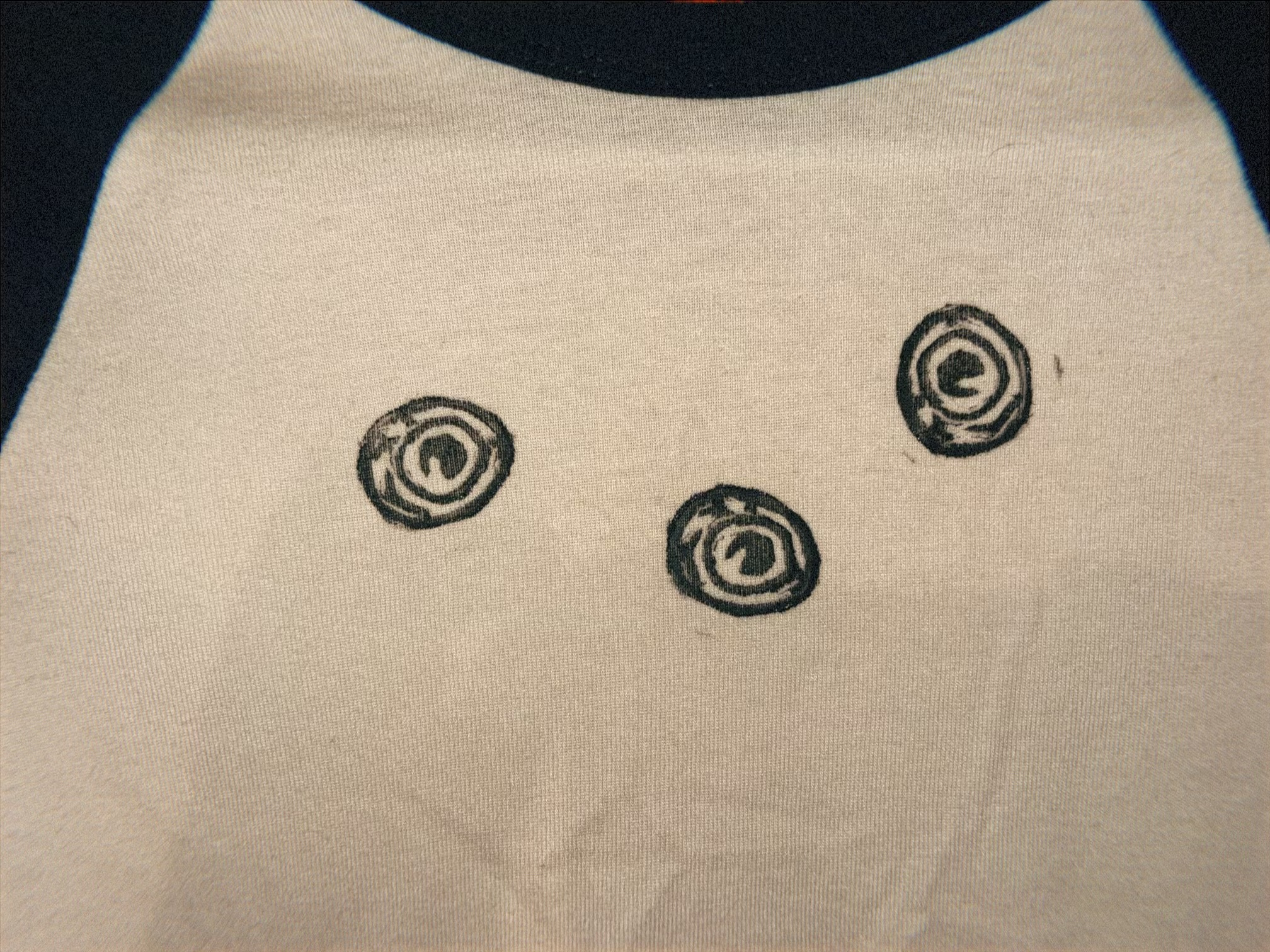 A close-up of a light beige fabric surface with three small black spiral or eye-like printed motifs arranged vertically. A dark curved shape appears along the right edge.