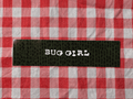 A close-up of a black textured fabric label with the words 'BUG GIRL' printed vertically in white, placed on a red and white checkered fabric background.