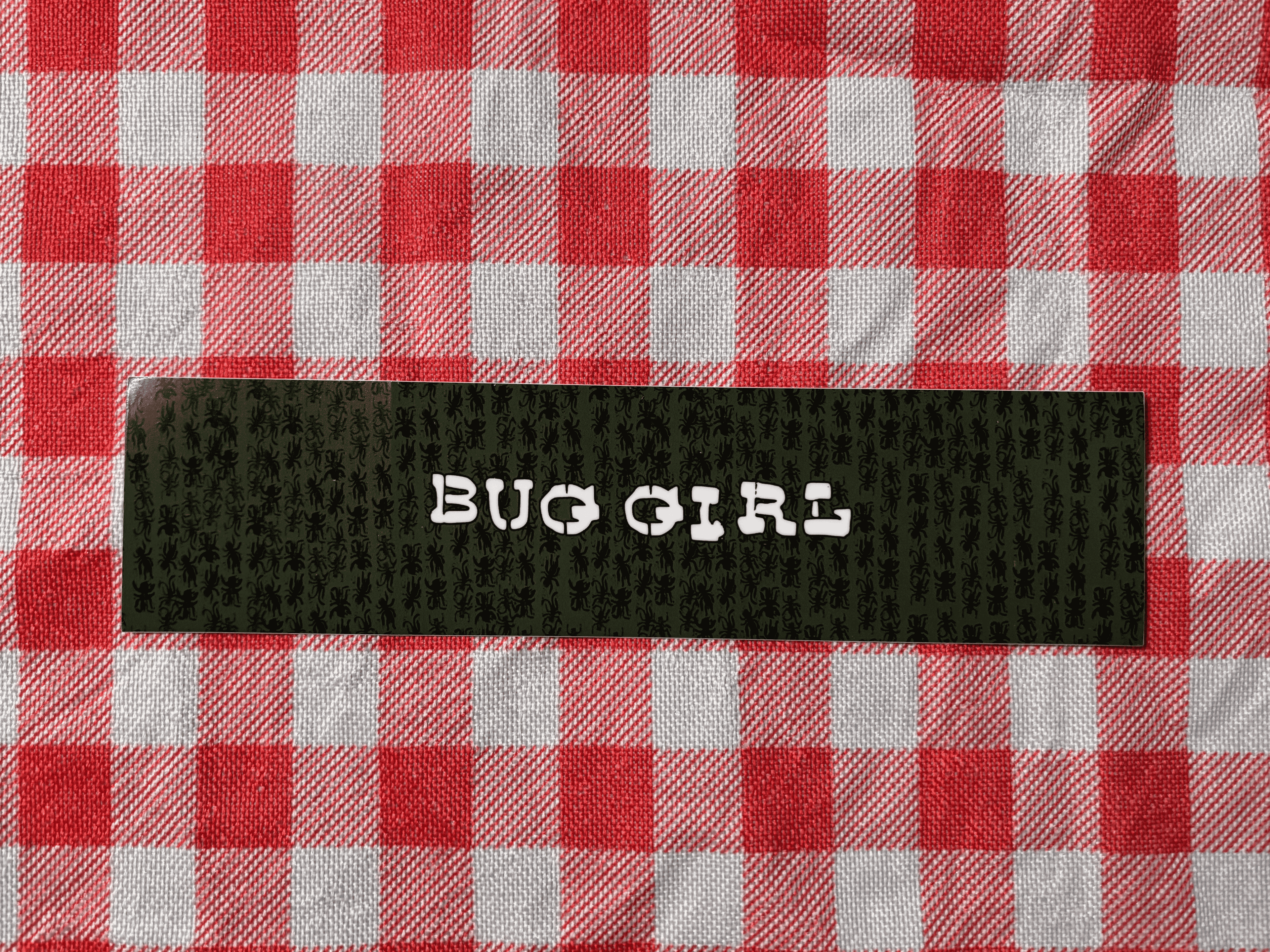 A close-up of a black textured fabric label with the words 'BUG GIRL' printed vertically in white, placed on a red and white checkered fabric background.