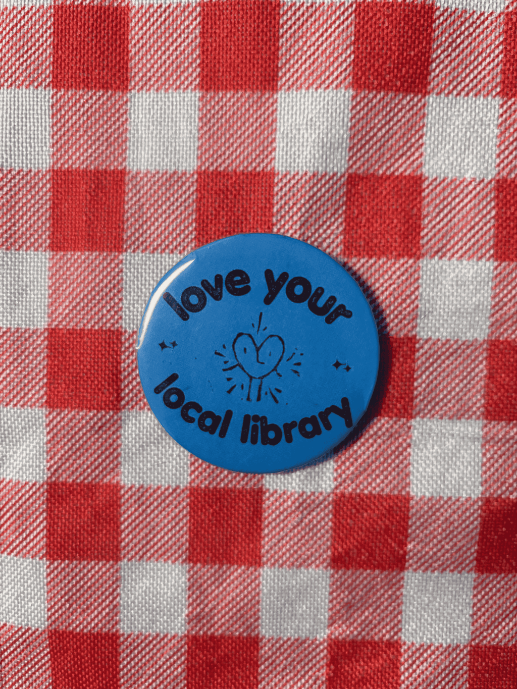 A round blue button pin with the text 'love your local library' and a simple heart illustration, placed on a red and white checkered fabric background.