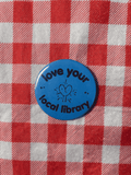 A round blue button pin with the text 'love your local library' and a simple heart illustration, placed on a red and white checkered fabric background.