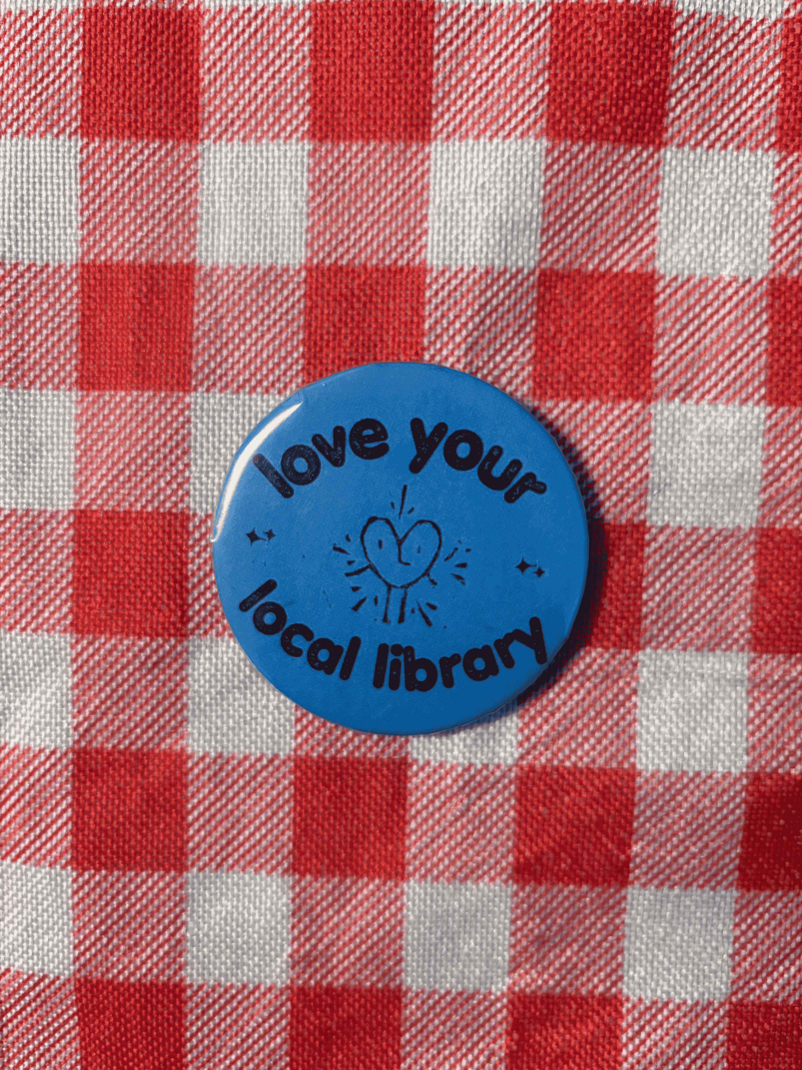 A round blue button pin with the text 'love your local library' and a simple heart illustration, placed on a red and white checkered fabric background.