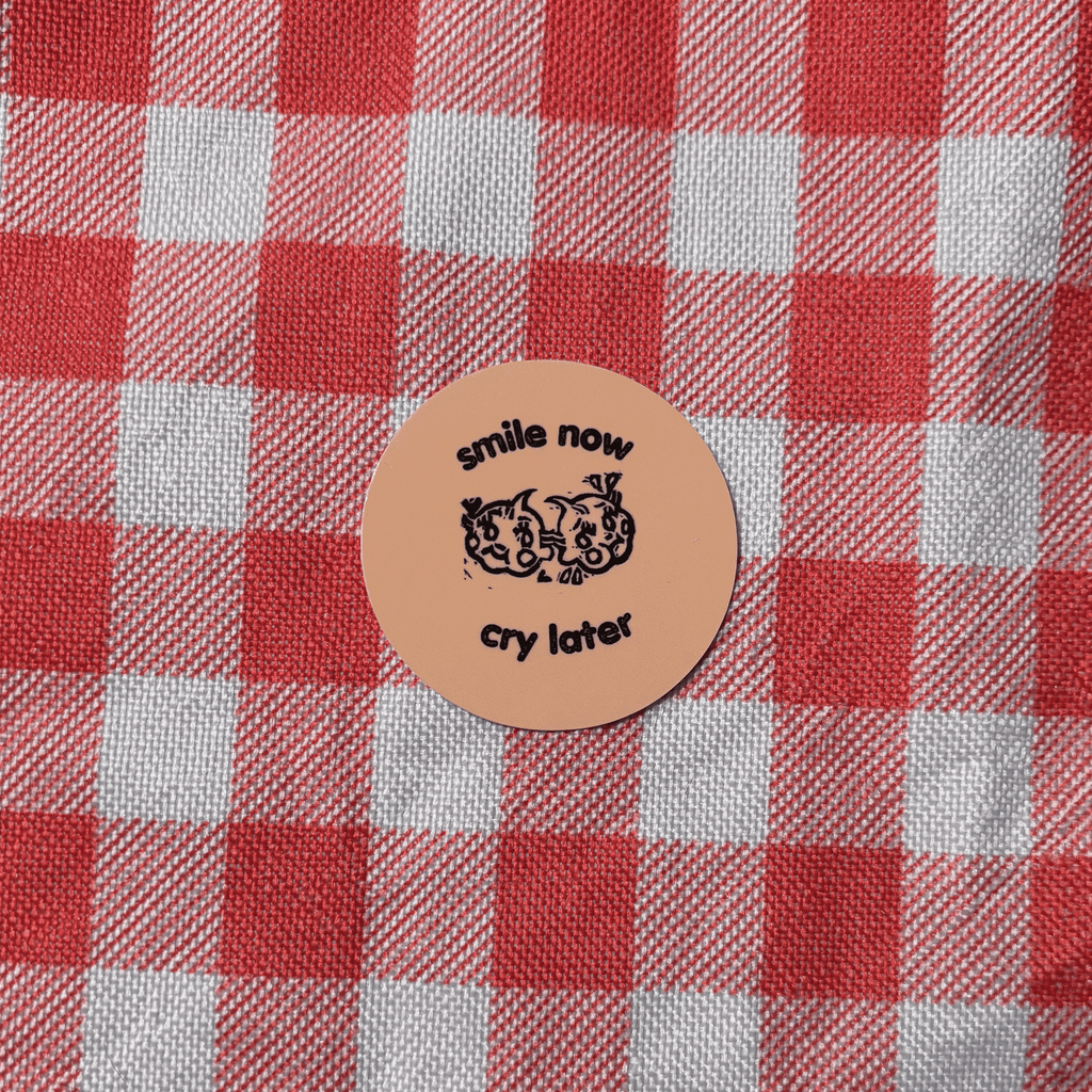 A circular tan sticker with black text reading 'smile now cry later' and a small graphic, placed on red and white checkered fabric.