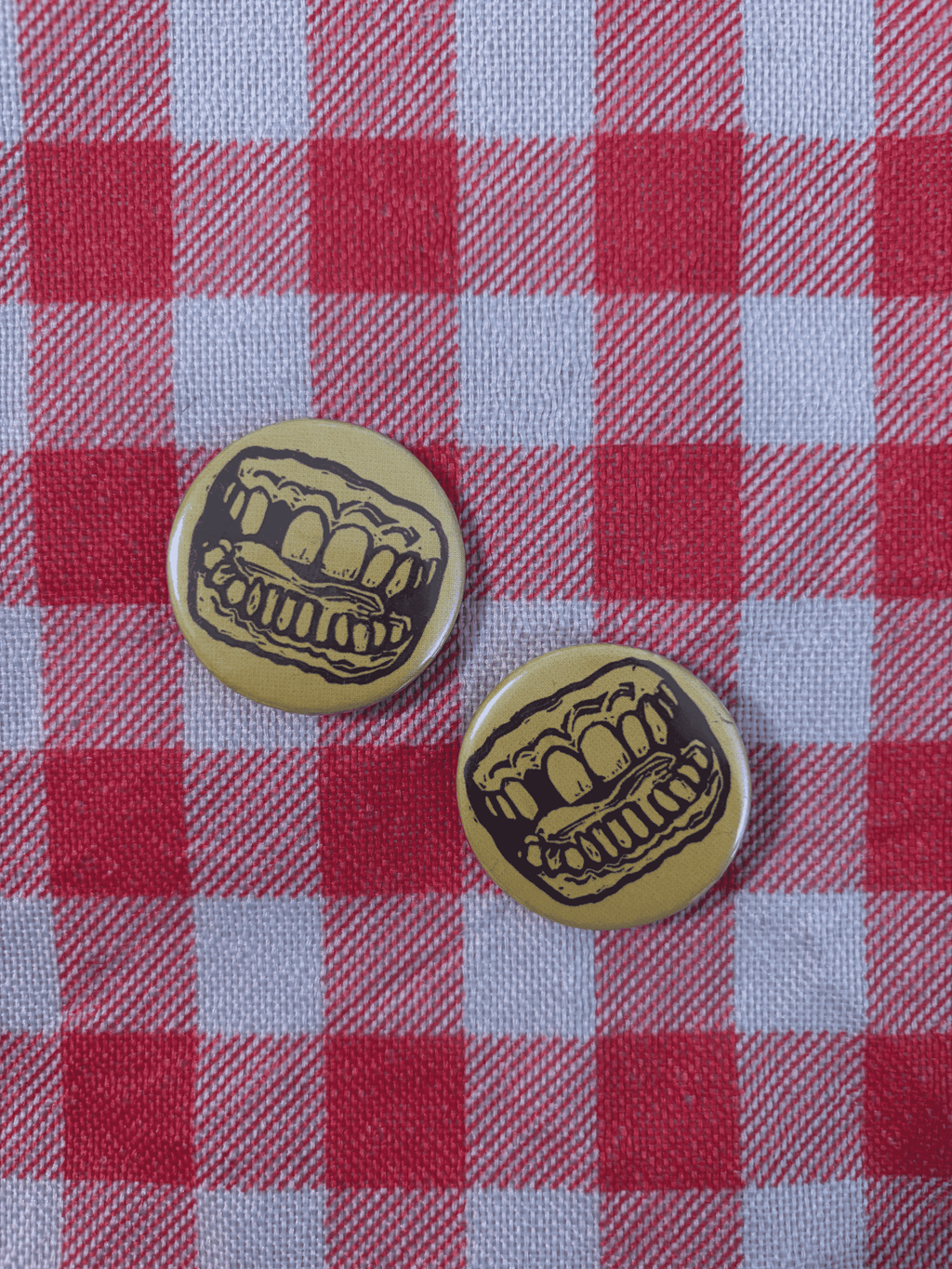 Two round pinback buttons featuring a black cartoon mouth with teeth on a yellow background, placed on a red and white checkered picnic blanket.