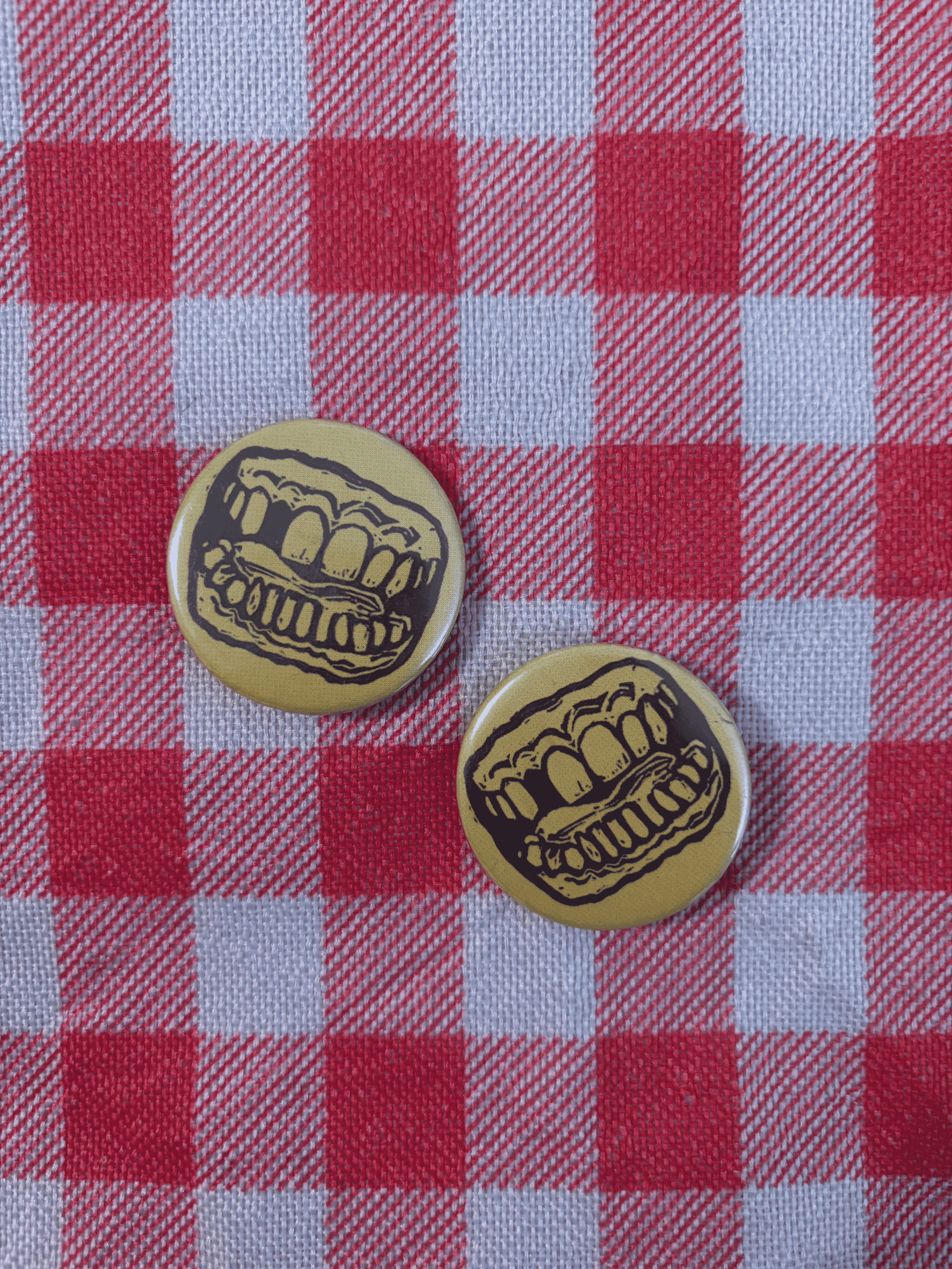 Two round pinback buttons featuring a black cartoon mouth with teeth on a yellow background, placed on a red and white checkered picnic blanket.