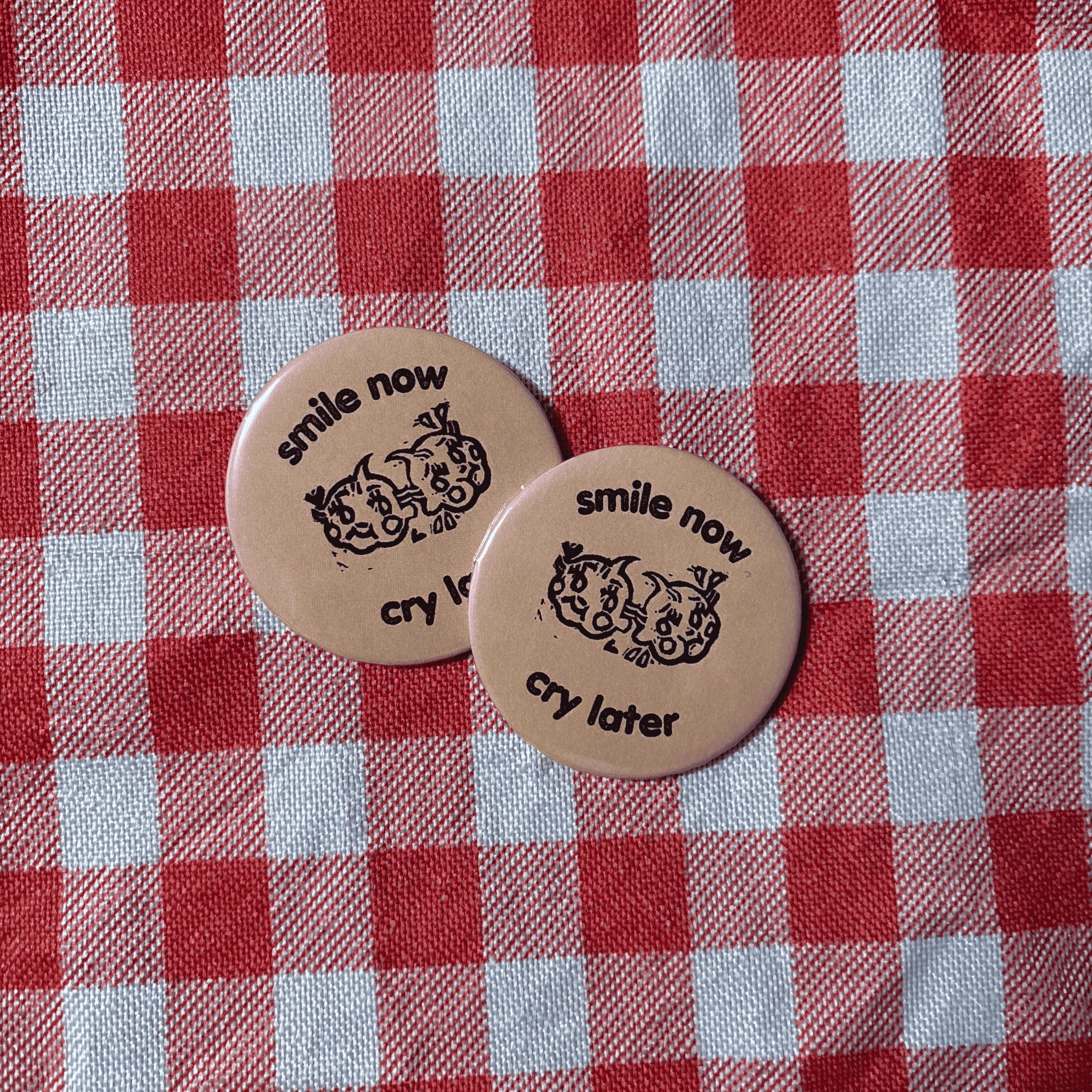Two round beige pin-back buttons with black text reading 'smile now cry later' and a cartoon face, placed on a red and white checkered fabric background.